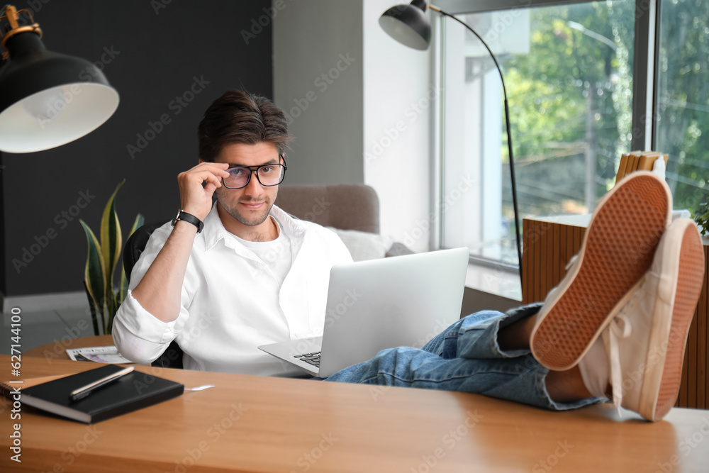 Handsome young man with wristwatch  and eyeglasses working on laptop having put his legs on table in