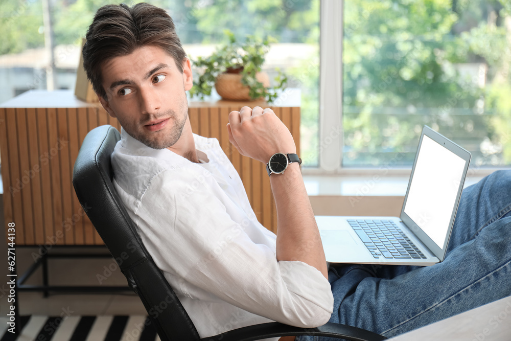 Handsome young man with wristwatch working on laptop having put his legs on table in office