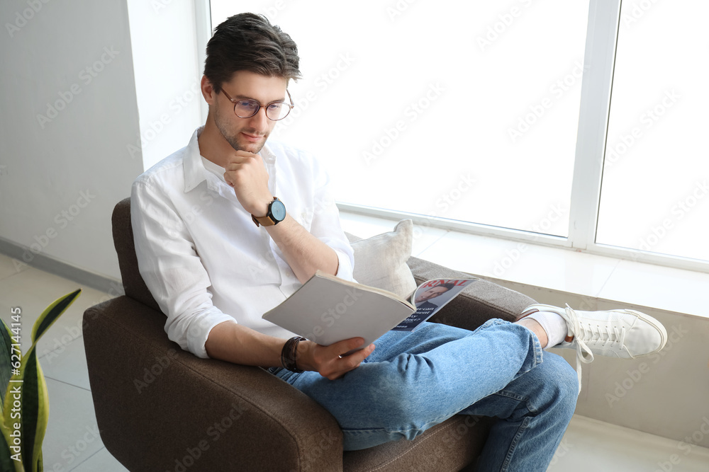 Handsome young man with wristwatch and eyeglasses sitting on armchair and reading magazine in room