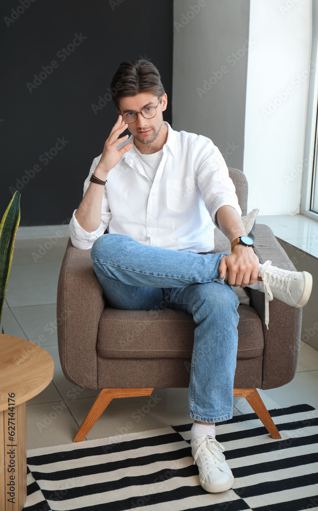 Handsome young man with wristwatch and eyeglasses sitting on armchair in room