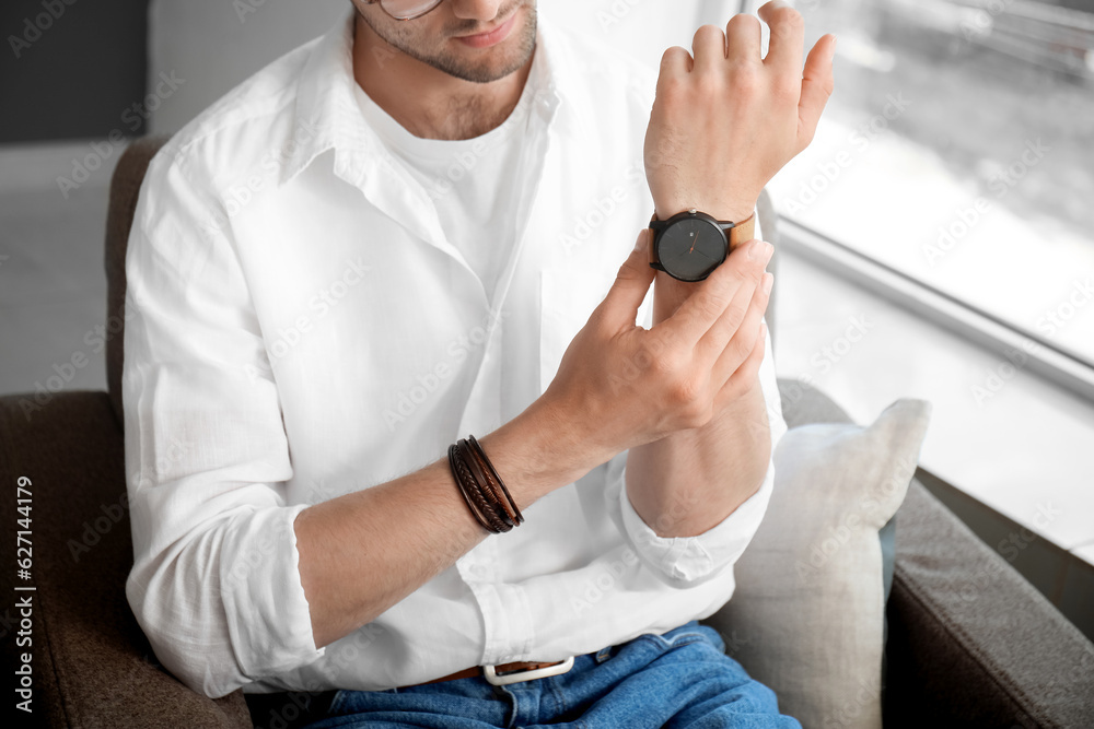 Stylish young man sitting on armchair looking at wristwatch in room