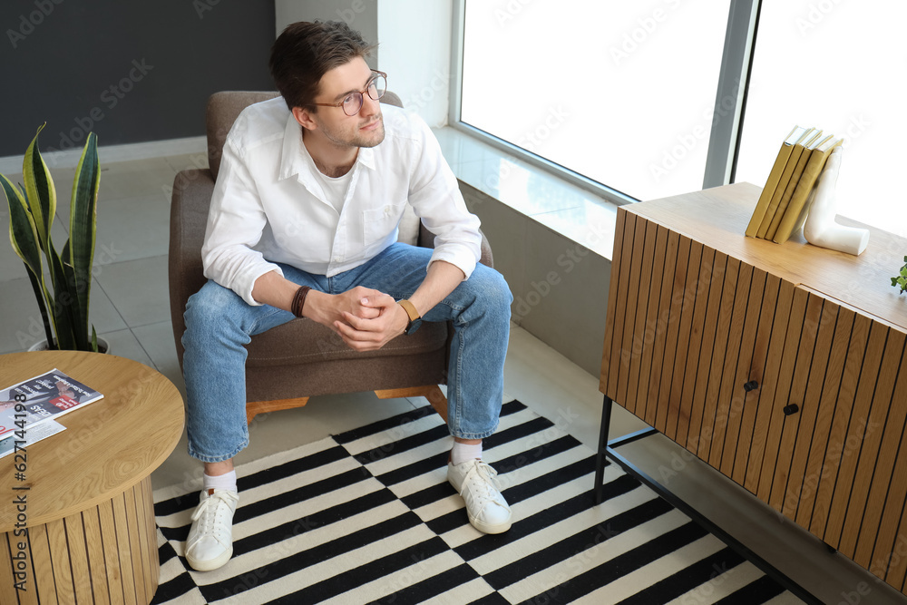 Handsome young man with wristwatch and eyeglasses sitting on armchair in room