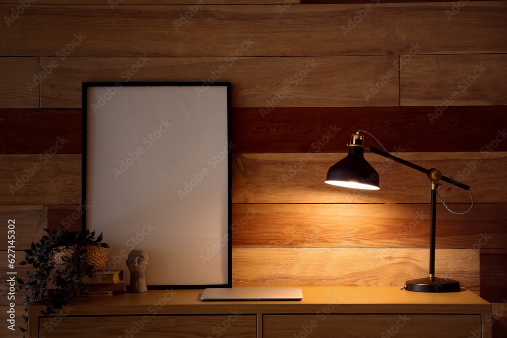 Desk with glowing lamp, modern laptop, blank frame and houseplant near wooden wall