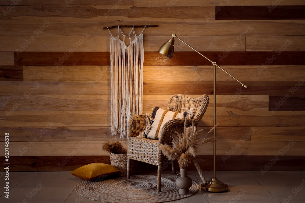 Cozy wicker armchair, glowing lamp and macrame near wooden wall