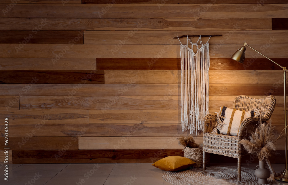 Cozy wicker armchair, glowing lamp and macrame near wooden wall