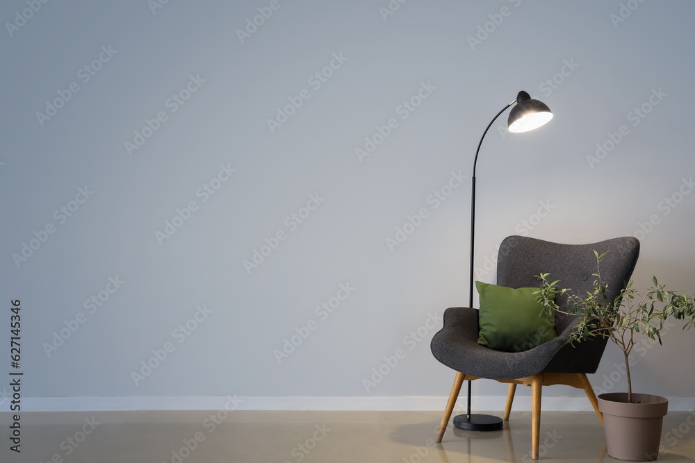 Glowing lamp, grey armchair with cushion and houseplant near grey wall