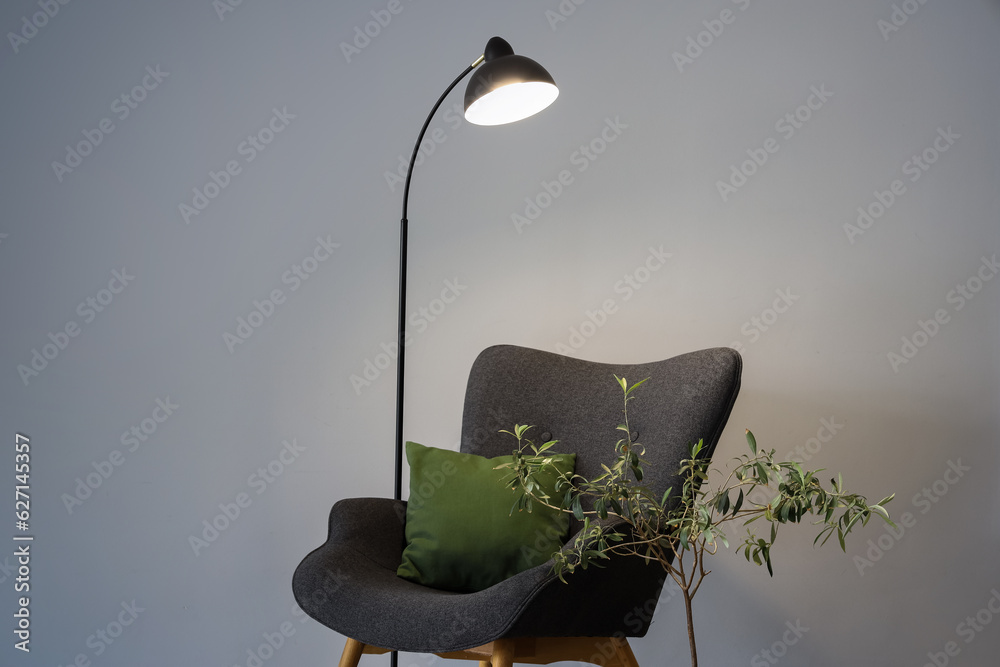 Glowing lamp, grey armchair with cushion and houseplant near grey wall