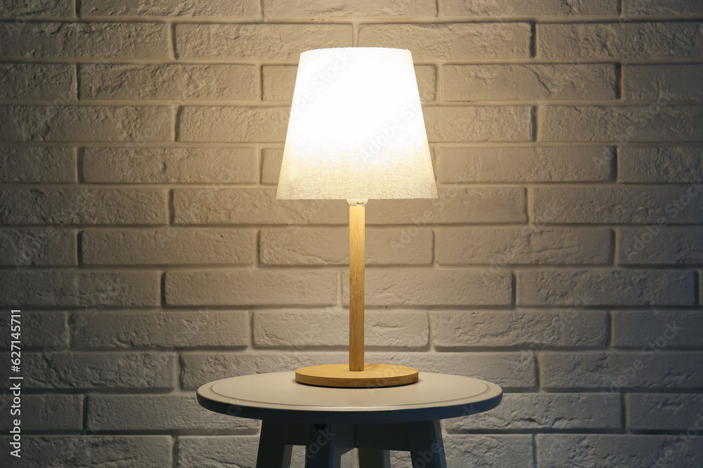 Glowing lamp on wooden table near white brick wall in dark room