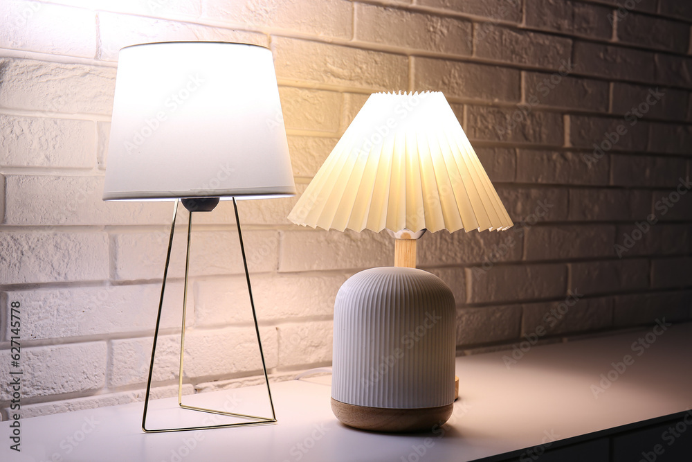 Different glowing lamps on table near white brick wall in dark room