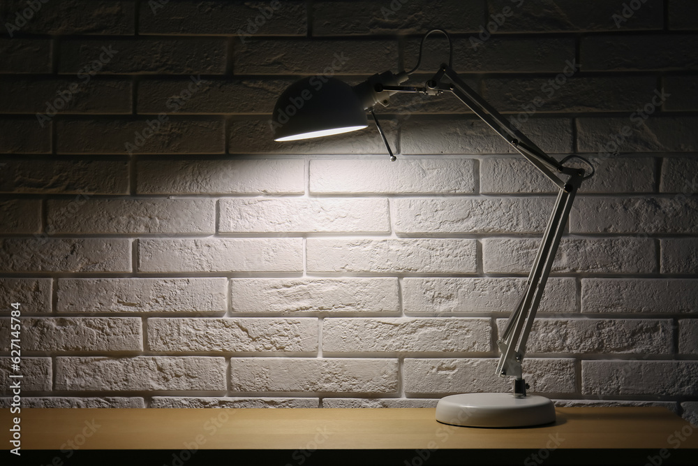 Glowing lamp on wooden table near white brick wall in dark room