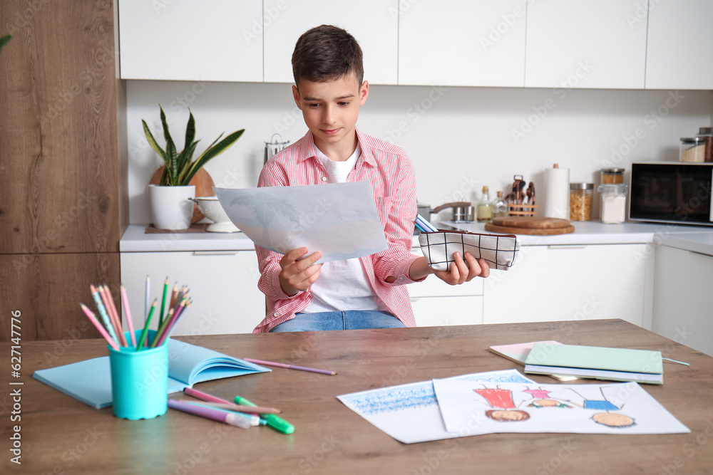 Little boy with pencil case and drawing at table in kitchen