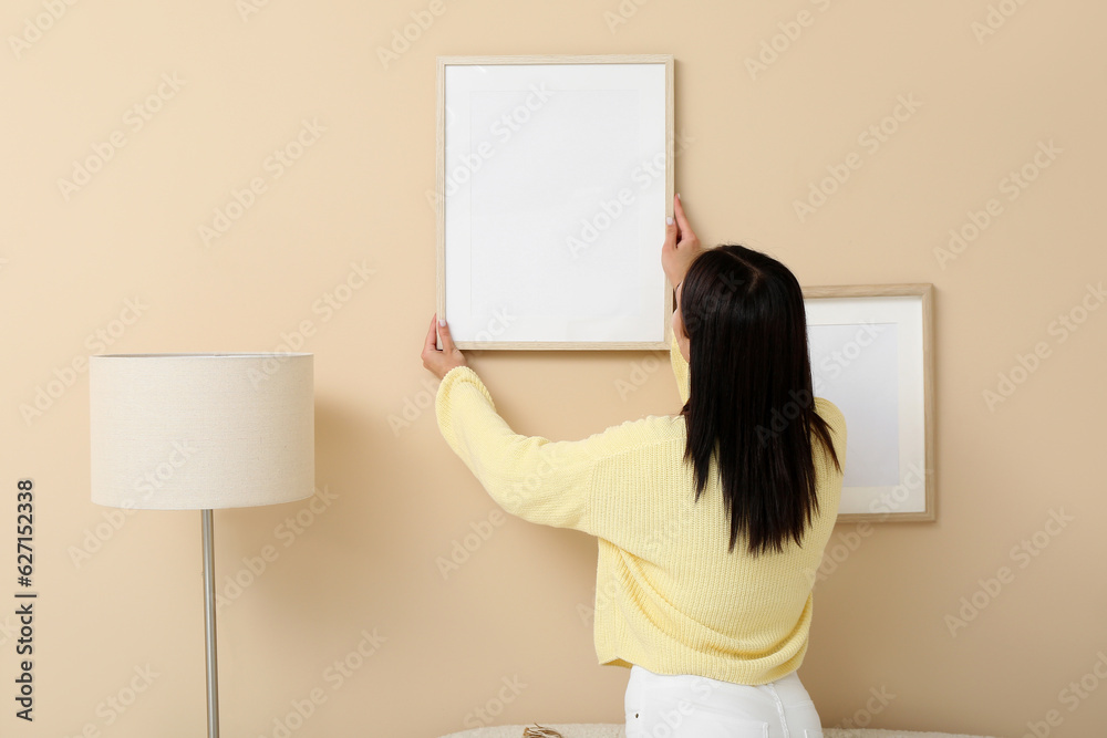 Young woman hanging blank frame on beige wall in living room, back view