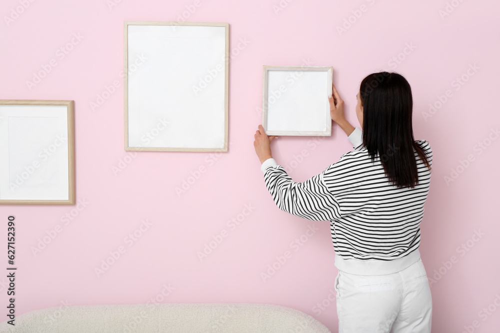 Young woman hanging blank frame on pink wall in living room, back view