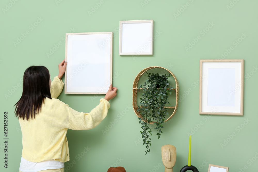 Young woman hanging blank frame on green wall at home, back view