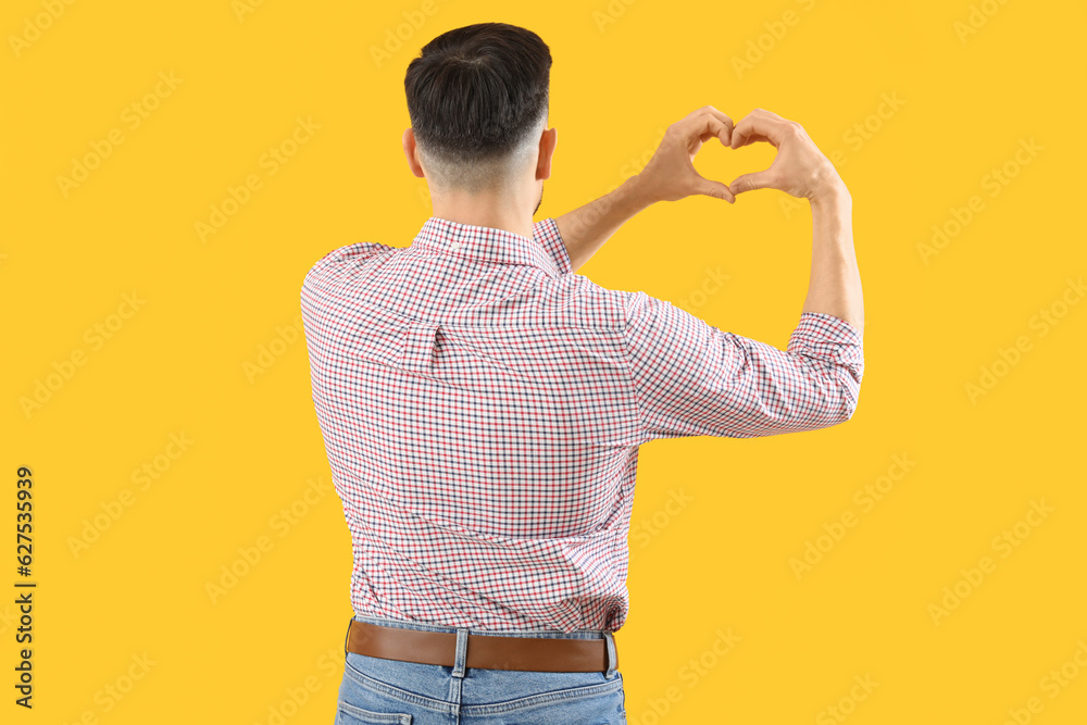 Handsome man making heart with his hands on yellow background, back view