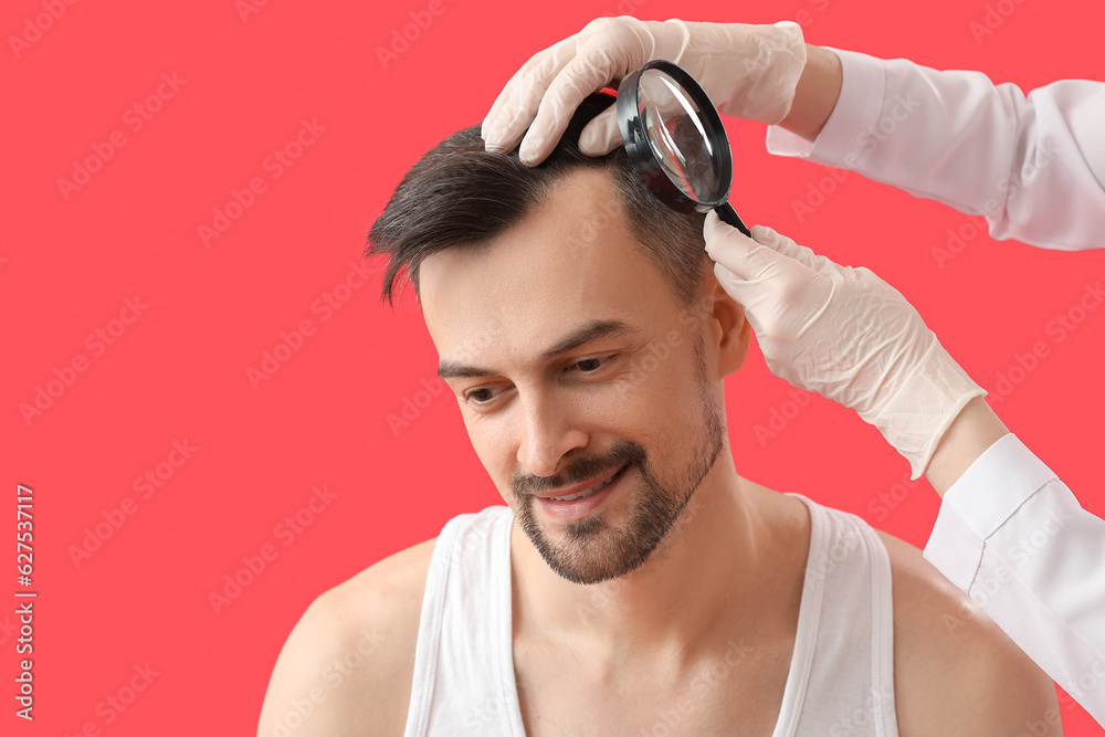 Doctor examining young mans hair with magnifier on red background, closeup