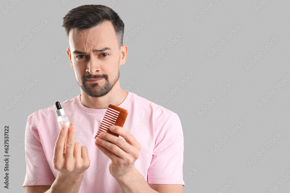 Worried young man with comb and serum for hair growth on grey background