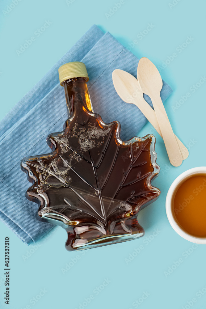 Bottle with saucer of tasty maple syrup on blue background