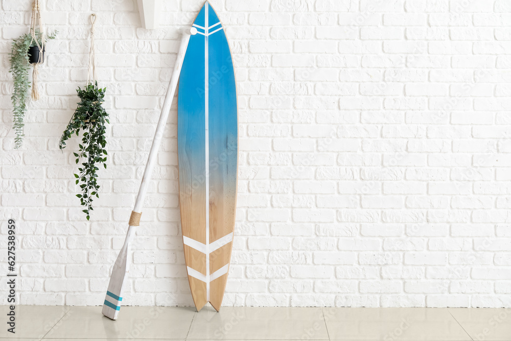 Wooden surfboard with paddle and houseplants on white brick wall in room