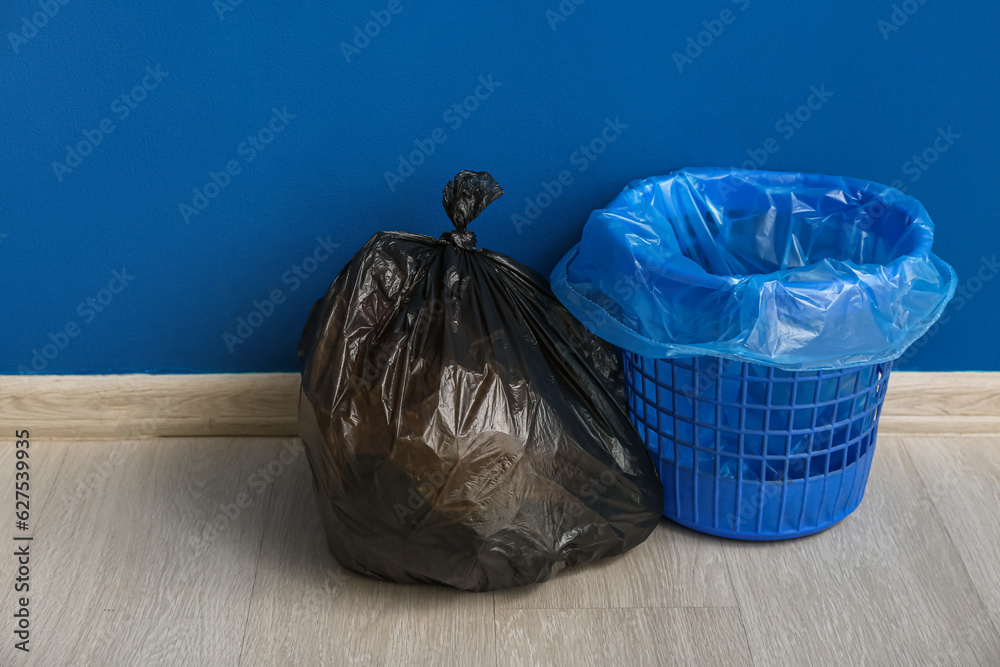 Full garbage bag and empty trash bin near blue wall