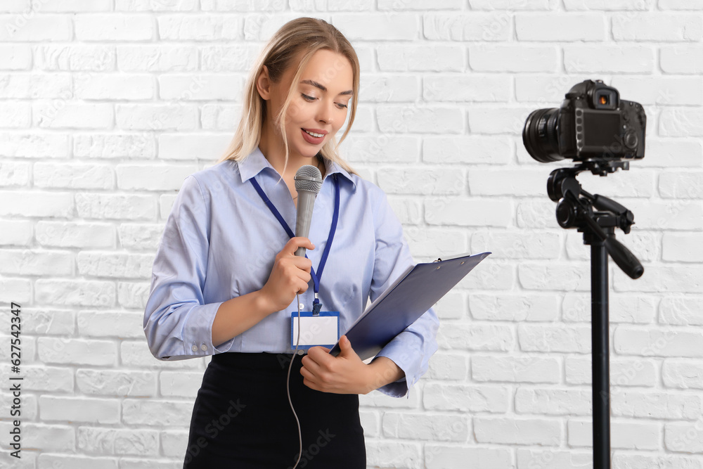 Female journalist with microphone and clipboard recording video on white brick background