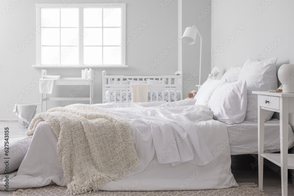 Interior of light bedroom with bed, changing table and baby crib