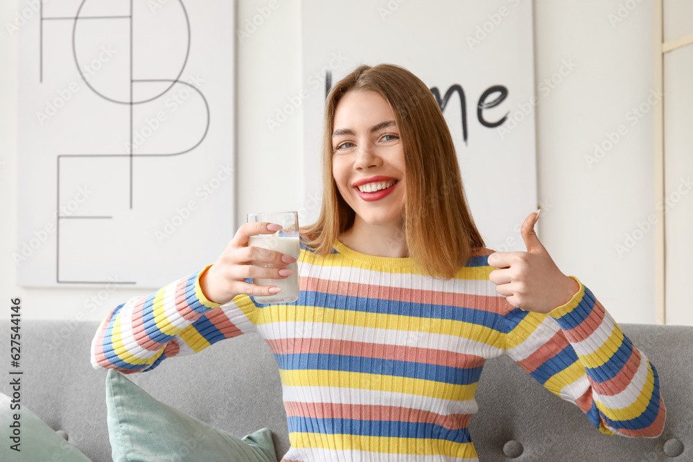 Young woman with glass of milk showing thumb-up at home