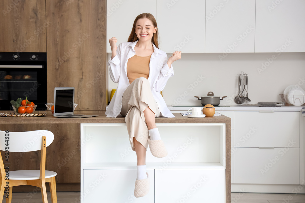 Happy young woman sitting on table with modern laptop in light kitchen