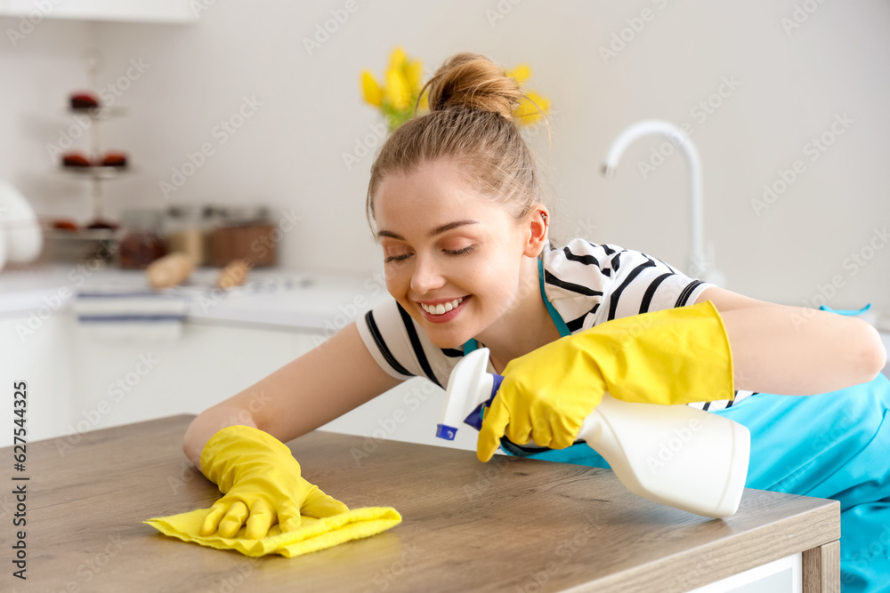 Happy young woman in yellow rubber gloves cleaning dining table with rag and detergent