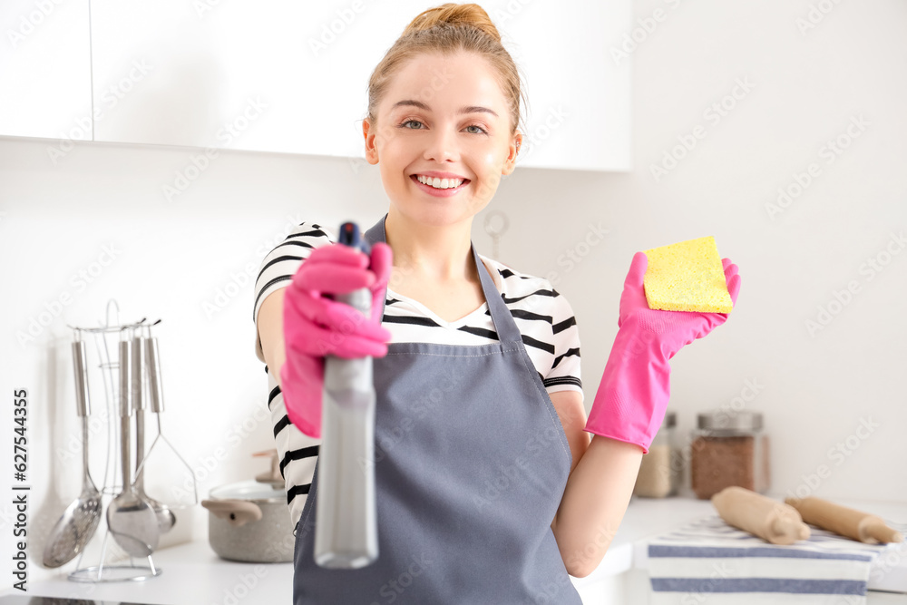 Happy young woman in rubber gloves with sponge and bottle of detergent in light kitchen