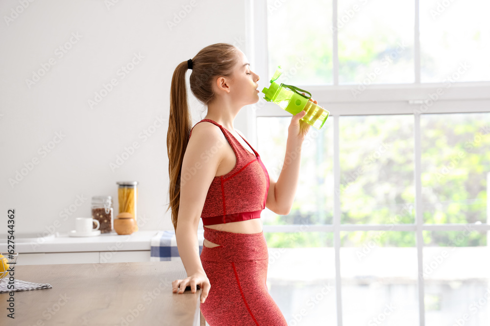 Sporty young woman drinking water from bottle in light kitchen