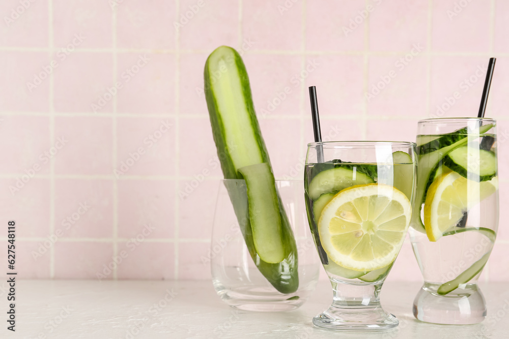 Glasses of lemonade with cucumber on white table near pink tile wall