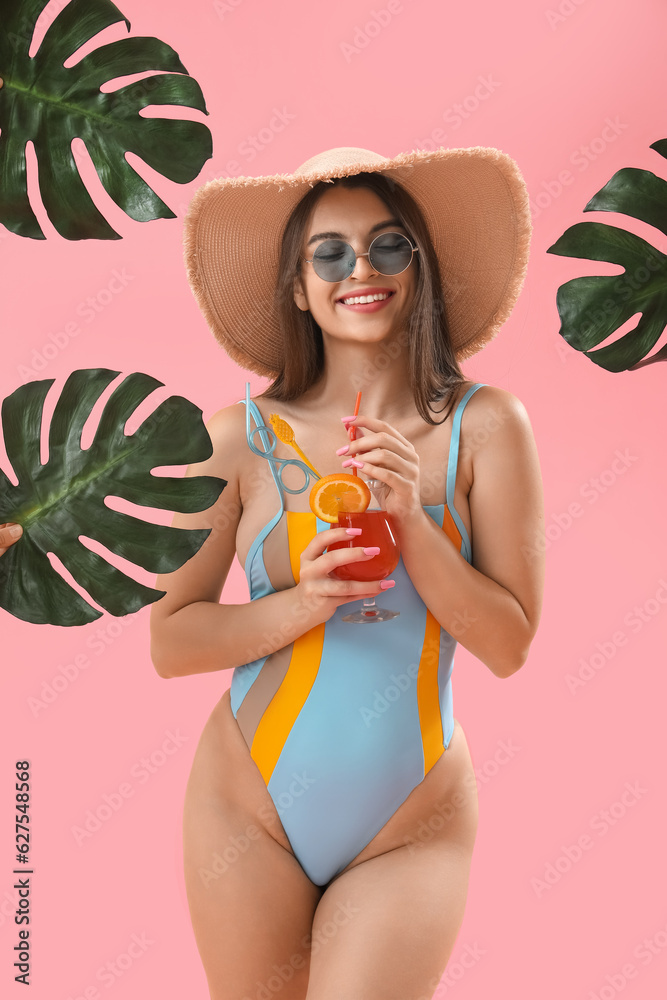 Young woman with palm leaves drinking summer cocktail on pink background