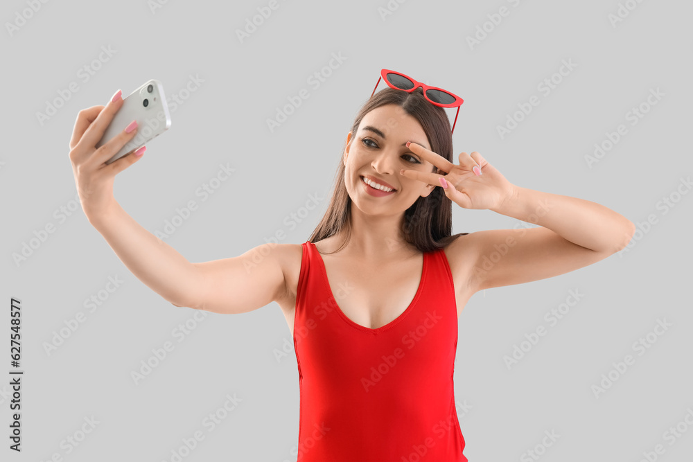 Young woman in swimsuit with mobile phone taking selfie on light background