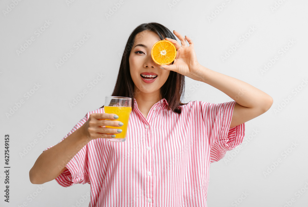 Beautiful Asian woman with orange and glass of juice on light background