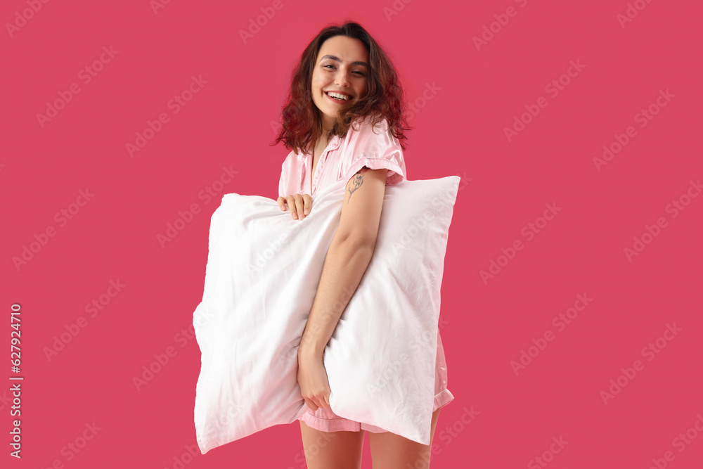 Young brunette woman in pajamas with pillow on pink background