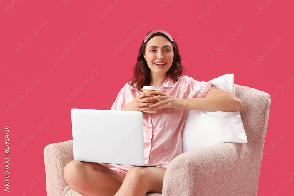 Young woman in pajamas with cup of coffee using laptop in armchair on pink background