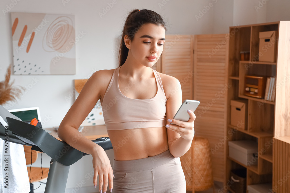 Sporty young woman using mobile phone near treadmill at home