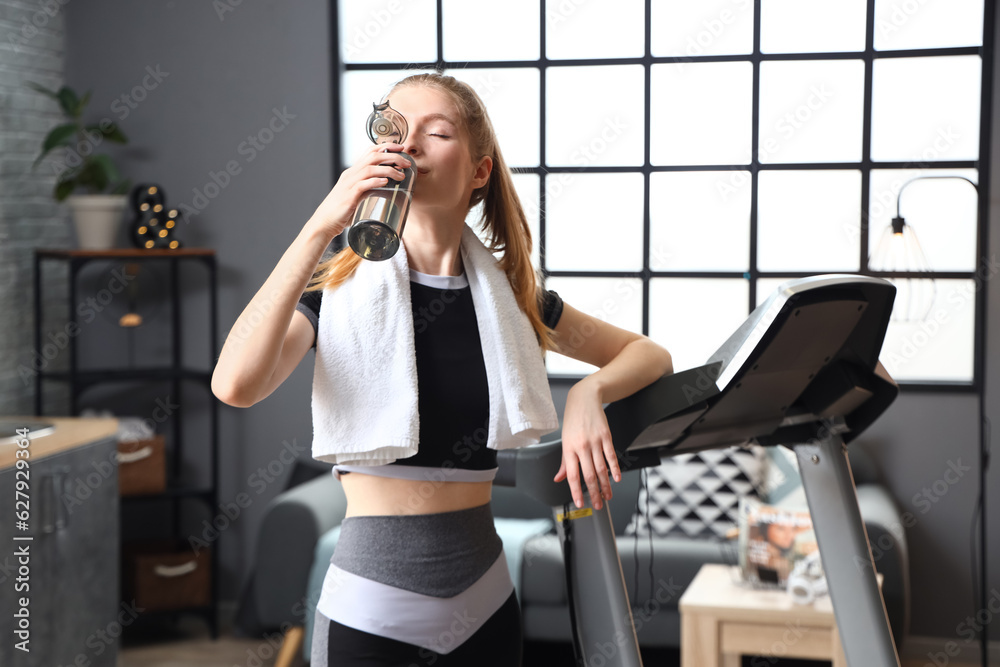 Sporty young woman drinking water after training on treadmill at home