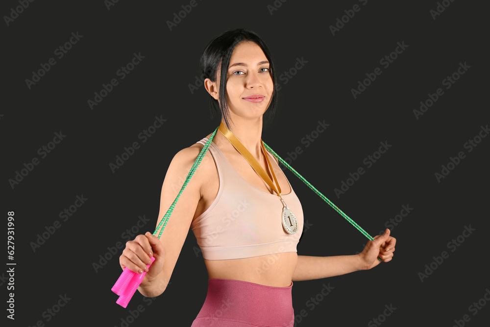 Sporty young woman with skipping rope and first place medal on black background