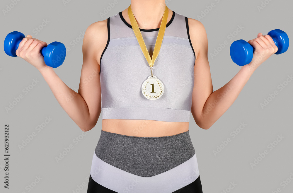 Sporty young woman with first place medal and dumbbells on grey background, closeup