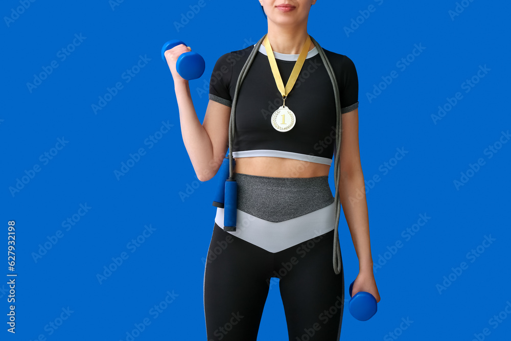 Sporty young woman with first place medal, skipping rope and dumbbells on blue background