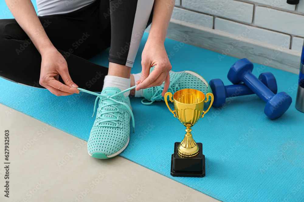 Sporty young woman with gold cup tying shoe laces in gym, closeup