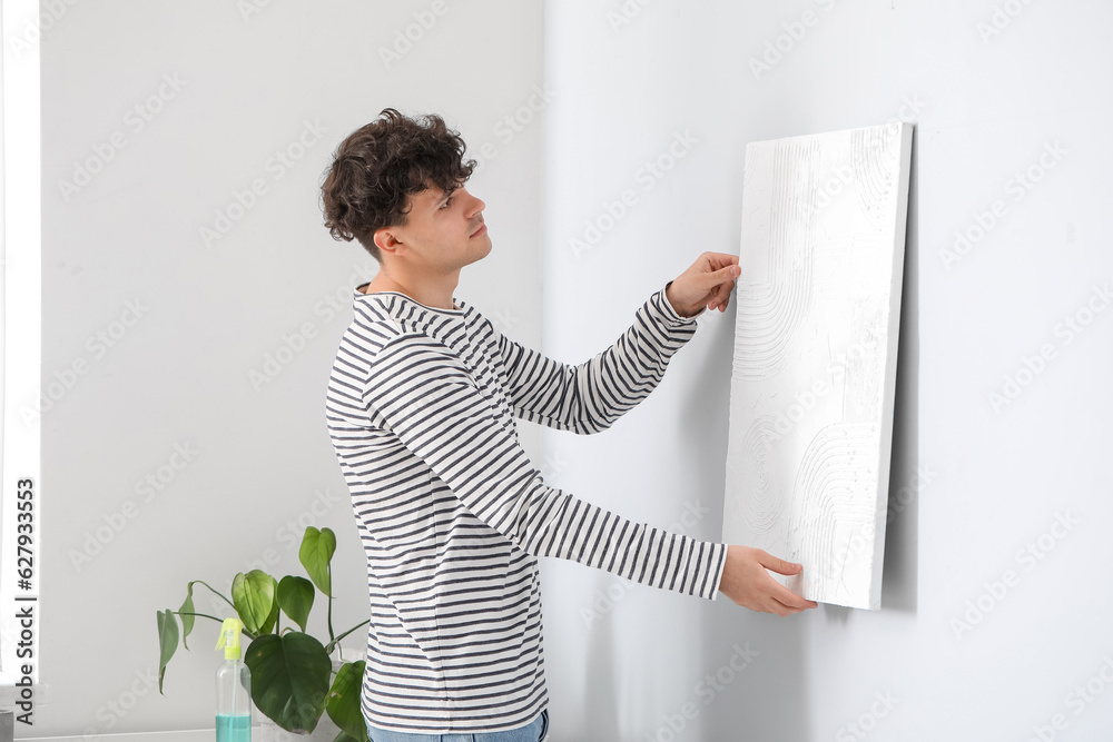 Young man hanging painting on light wall in bedroom