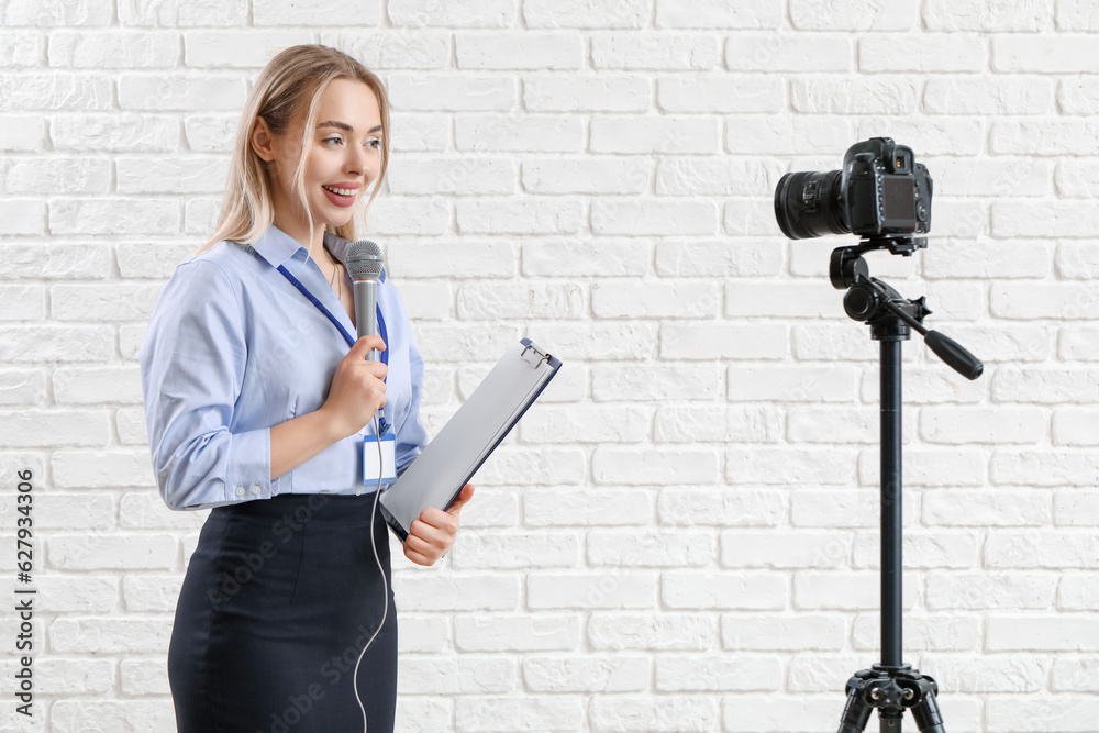 Female journalist with microphone and clipboard recording video on white brick background