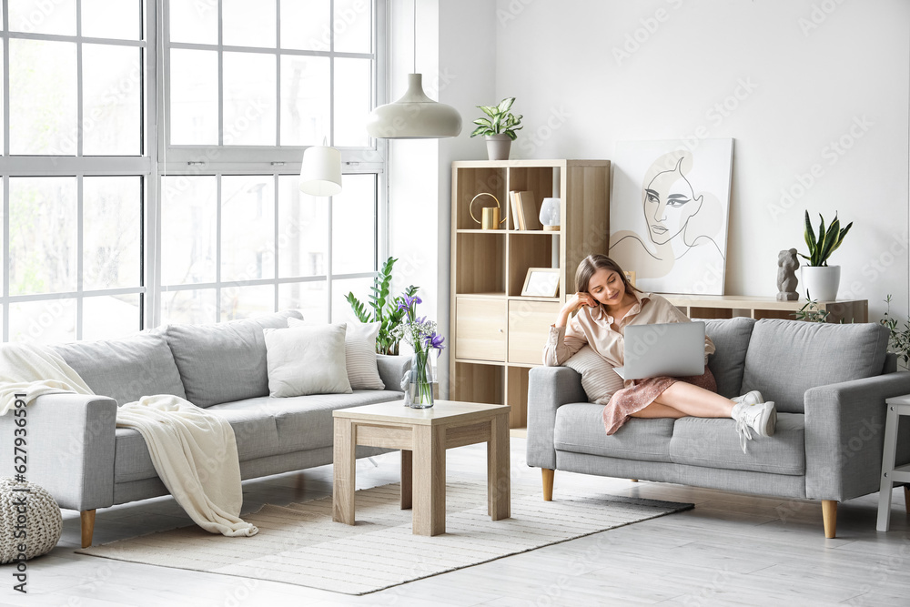 Pretty young woman sitting on grey sofa and using laptop in light living room