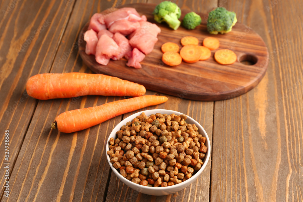 Bowl with dry pet food, raw meat and natural products on wooden background