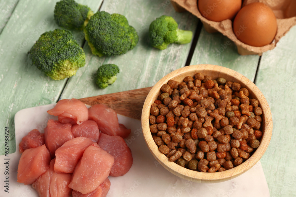 Bowl with dry pet food, raw meat, eggs and broccoli on color wooden background, closeup