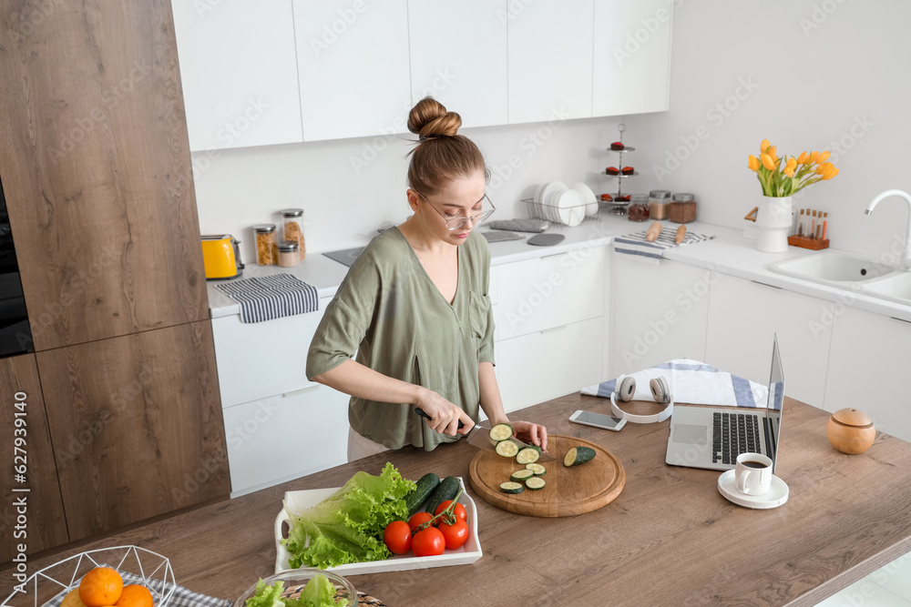 Beautiful young woman cutting cucumber at table with modern laptop in light kitchen