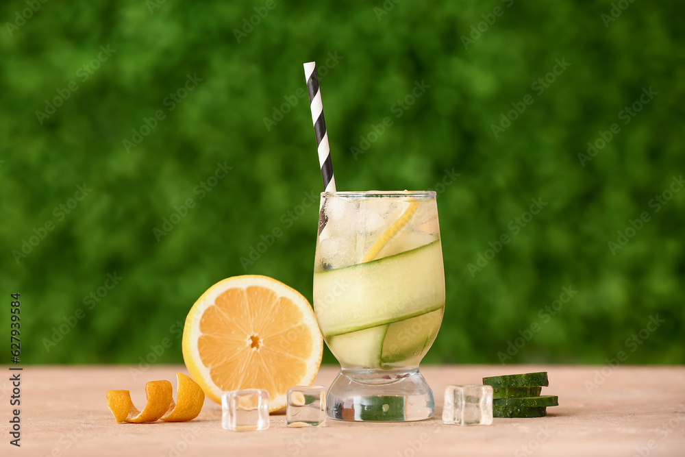 Glass of lemonade with cucumber and ice cubes on beige table outdoors